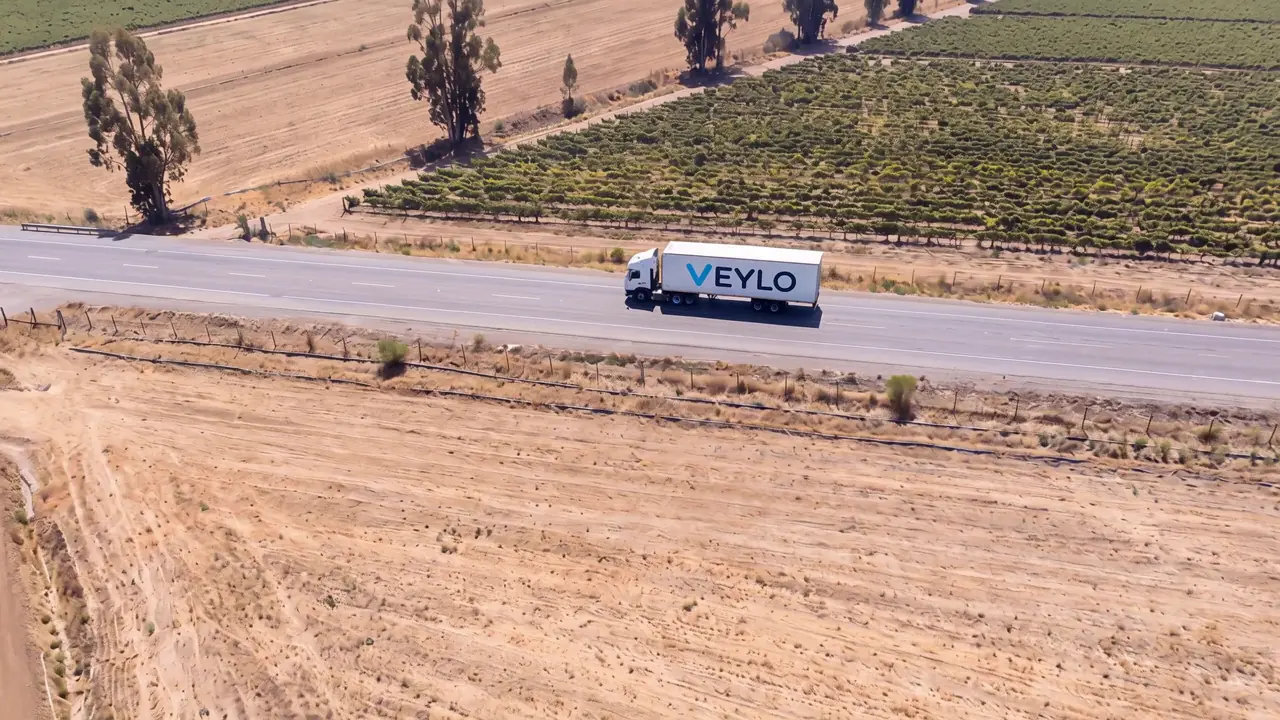 Camión de Veylo avanzando por una carretera del valle central entre campos de cultivo, visto desde el aire.
